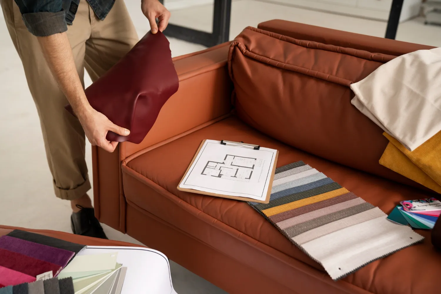 Persona ajustando un cojín rojo en un sofá marrón, con muestras de colores y un plano de diseño sobre la mesa. Ideal para decoración de interiores.